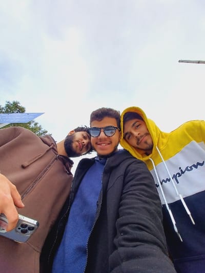 The image displays three young adult males captured from a low-angle perspective against an overcast sky, in what is identified as Al-Muhajerin Municipality, Syria.
The individual on the far left, partially visible, wears a brown zippered hooded sweatshirt. Their right hand, holding a silver smartphone with a multi-lens camera array, is extended into the lower left foreground, indicating they are operating the camera. This subject has a beard and looks directly at the camera with a neutral expression.
The central figure, fully visible from the shoulders up, wears a black jacket over a blue garment. Dark sunglasses obscure their eyes. This individual has dark, curly hair, visible dental braces, and is smiling towards the camera.
The individual on the far right, partially visible, wears a yellow, white, and dark blue color-block hooded sweatshirt with "Champion" lettering and white drawstrings. This subject has facial hair and exhibits a slight smile while looking towards the camera.
The background primarily consists of a uniformly light grey, overcast sky. To the far left, a portion of a blue solar panel array is visible, situated near green foliage. To the far right, a thin, light-colored pole extends vertically into the upper right corner of the frame. The scene suggests an outdoor, informal gathering or selfie.