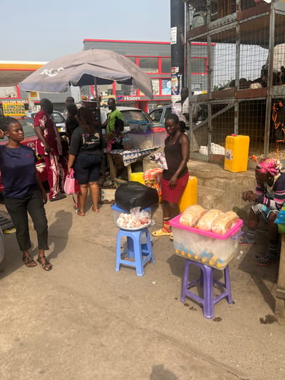 A street scene displays several vendors selling goods. One table has eggs and bread for sale, another vendor is selling something off of a yellow container. Customers are lined up in front of the sales areas. In the background is a building with a large Haier advertisement.