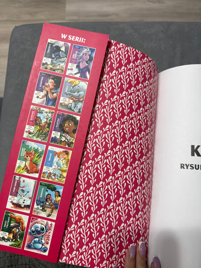 The image shows a close-up, top-down view of a red booklet featuring various Disney characters. The booklet is patterned with white leaves on a pink background. The cover displays multiple rectangular cards, each with a different Disney character illustration and text. Some of the characters visible include Elsa from Frozen, Dumbo, Stitch, Snow White, Jasmine, Bambi, and characters from Lilo & Stitch and Beauty and the Beast. The text on the cards includes phrases like "GIRL POWER," "DZIECI" (children), "KLASYKA" (classic), "MIŁOŚĆ" (love), "RODZINA" (family), "KOLORY NATURY" (colors of nature), "ŚWIAT PRZYRODY" (world of nature), and "PORY ROKU" (seasons). The phrase "W SERII:" (in series:) is also visible on the left side of the booklet. A portion of a white paper with black text, including the letter "K" and "RYSU," is visible at the top. A hand with purple nail polish is seen at the top right corner, resting on the patterned booklet. The setting appears to be indoors, possibly on a sofa or similar surface.