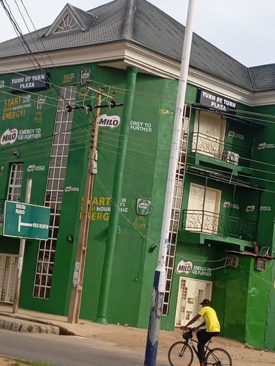 The image shows the exterior of a green building in Kabong, Nigeria. The building is adorned with multiple advertisements for Milo, a popular beverage. Some of the text visible on the building includes "TURN BY TURN PLAZA," "START WITH NOURISHING ENERGY," and "ENERGY TO GO FURTHER." There is a traffic sign on the left side of the image indicating directions to "RUKUBA MIANGO" and "ROCK HEAVEN." A man is seen riding a bicycle on the road in front of the building, wearing a yellow t-shirt and a black cap. Power lines and a utility pole are also prominent in the scene. The building appears to be a commercial establishment.