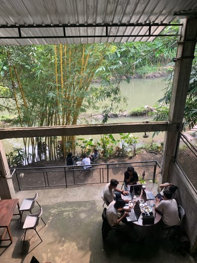 A group of five young adults sit around a round table on a concrete patio, working on laptops. The patio is enclosed by a black metal railing. Beyond the railing, a small river runs through a lush, green landscape. The view of the river is from an upper level, through a covered open area with a corrugated metal roof. There are bamboo plants growing along the river bank. The scene is located in Kecamatan Piyungan, Indonesia, at latitude -7.825, longitude 110.453.