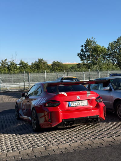 The image shows the rear of a red BMW M2 Competition parked in an outdoor setting. The car has a license plate that reads "AW TS 799". It is parked on a patterned paving surface, and to the left of the car is a metal fence. Beyond the fence, there are trees and a field, suggesting a rural or semi-rural location. The sky is clear and blue, indicating a sunny day. The angle of the sun suggests it is mid-day. The scene appears to be at a car track or a car event, given the presence of multiple sports cars and the racing-style spoiler on the red BMW. The overall mood is bright and energetic, consistent with a car enthusiast gathering. The location is Meuspath, Germany.