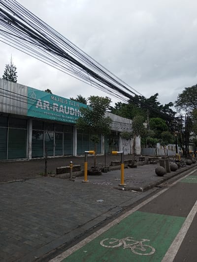 The image displays a quiet urban street scene under an overcast sky. A prominent single-story building occupies the left side of the frame, featuring multiple glass storefronts or windows. A large teal-colored banner sign spans the building's facade, displaying the text "MAJELIS TA'LIM AR-RAUDHAH," with smaller text beneath reading "PONDOK PESANTREN TAHFIDZ & JAME" and "JL. JETISAN NO.18 CITEUREUP CIMAHI." This textual information indicates the location is Cimahi, Indonesia. The requested location of Vaatharawattai, Sri Lanka, does not align with the visible textual evidence.
In front of the building, a grey paved sidewalk runs horizontally, lined with several small, sparsely leafed trees planted in recessed areas. A series of yellow and grey bollards or parking barriers are installed along the edge of this paved area. Further down the street, spherical concrete or stone barriers and decorative lampposts are visible along the roadside.
To the right of the sidewalk, an asphalt road features a clearly marked bicycle lane, painted green with a white bicycle symbol. Overlapping the upper section of the building and the sky, a dense bundle of black electrical or communication cables stretches diagonally across the frame. No individuals are present in the scene, and no actions or interactions are depicted. Denser foliage from larger trees is visible in the background, behind the building and along the street.