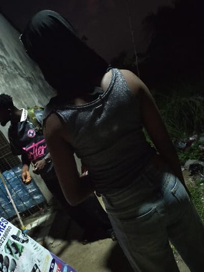 A person in a silver top and light jeans stands with their back to the camera. Their dark hair partially obscures their face. A man wearing a black and pink "Cartier" shirt is to the left, holding a small object. They are positioned in front of a building and a caged area filled with blue containers. The scene appears to be at night, with limited lighting, possibly outdoors near some vegetation and trash.