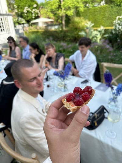 The image shows a hand holding a raspberry tart in focus, with a blurry scene of people seated around a table in the background. The table is covered with a white tablecloth and has floral arrangements. People are dressed in casual attire. The background suggests an outdoor setting with greenery and possibly a building. While it's difficult to confirm the exact location is Cambridge, UK without more information, the general aesthetic and ambiance could align with garden party settings common in Cambridge. More specific location details are not discernible from this image.