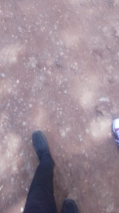 The scene, located in Kikuyu Township ward, Kenya, depicts an overhead, significantly blurred view of an unpaved outdoor ground surface and the lower extremities of two individuals. The ground is composed of reddish-brown soil interspersed with scattered lighter-colored gravel or small stones. On the lower-left, a person's left leg is visible, clad in dark trousers and a dark, closed-toe shoe. On the lower-right, the toe and partial side of a second person's right shoe are visible, appearing to be a light-colored sneaker with darker accents. Both individuals are positioned on the unpaved ground, their exact actions or interactions indiscernible due to the image's lack of focus.