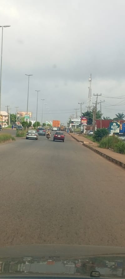 The image displays a daytime street scene on a multi-lane asphalt road in Akure, Nigeria, under an overcast sky. The perspective is from inside a vehicle, indicated by a dashboard reflection across the bottom edge.
Traffic is moving forward in the visible lanes. In the left lane, a silver sedan with an open trunk, containing white items, is followed by a dark blue or grey sedan. A yellow and black motorcycle with two occupants is positioned between the lanes. In the right lane, a red sedan is followed by a truck carrying a large orange shipping container. Behind this, a yellow construction vehicle is visible.
The left side of the road features a series of tall streetlights, power poles, and partially obscured buildings behind vegetation, with multiple billboards visible above.
The right side of the road includes additional power poles, a prominent telecommunication tower, and dense vegetation. Commercial establishments are present, characterized by multiple shipping containers, some dark red/brown and others blue. One blue container prominently displays "AYA SUPER HOTEL AKURE." Further billboards are visible. A single pedestrian in a blue outfit is observed walking on the far left sidewalk, away from the camera. Other indistinct figures are present on the right sidewalk. The asphalt road surface exhibits wear.