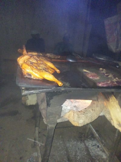 A whole roasted chicken sits on a grill next to raw beef. Under the grill, a wood fire burns in a metal structure. People stand in the background, though details are obscured by darkness and possible smoke. The lighting is dim.