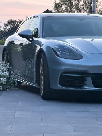 A gray Porsche Panamera is parked on a gray stone tiled driveway in Marche-en-Famenne, Belgium. The front of the car is closest to the camera, showing the distinctive headlight and air intake. A patch of daisies is visible near the car's rear wheel. The background shows some trees and sky, indicating an outdoor setting. The overall lighting is soft, suggesting either early morning or late evening.