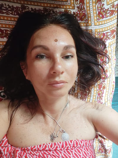 A woman lies on a patterned cloth in a room in Puttaparthi, India. The woman is lying on her back. She is wearing a red and white patterned top and a silver necklace. She has dark hair and green eyes. There is a golden bindi on her forehead. The patterned cloth she is lying on is predominantly yellow and white. The room appears to be lit by natural light.