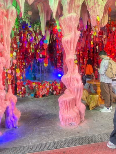 The image shows a cave-like structure decorated with pink stalactites and stalagmites, creating a whimsical and surreal atmosphere. The cave is filled with red and pink paper hearts, which are hanging from the ceiling, creating a sense of romance and tradition. In the center of the cave, a small fountain with blue water is surrounded by more paper hearts.
Two people are present in the scene. One person is standing on the right side of the image, wearing a plaid shirt and a backpack, looking towards the fountain. The other person is standing on the left side of the image, wearing an orange shirt and facing away from the camera. They appear to be interacting with a wooden table and stool, situated in front of the fountain. The scene is illuminated by a combination of natural light and artificial light, creating a soft and ethereal ambiance.
The location is a man-made cave structure, built as a part of a tourist attraction, located in Hang Zhou Shi, China, at coordinates (latitude: 30.48, longitude: 120.179).