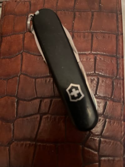 A black Swiss Army knife with a white cross emblem on the handle rests on a brown textured surface that resembles alligator skin. The knife is oriented diagonally, with the handle at the top left and the blade extending towards the bottom right. A silver keychain ring is attached to the end of the knife. The textured surface appears to be a notebook cover. The lighting in the image is dim, casting shadows on the textured surface and highlighting the glossy finish of the knife. There is no indication of the location being in Dzoraghbyur, Armenia.