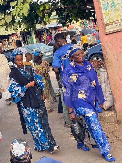 This image depicts a bustling street scene in Samaru, Nigeria, likely a market or commercial district, under daylight conditions. The ground is an unpaved, dusty surface.
In the left foreground, a young adult female is visible, facing slightly right and looking downwards. She wears a blue headwrap, a long-sleeved gown featuring a blue, black, and white abstract pattern, and a solid black shawl draped over her shoulders. A silver-colored bracelet adorns her left wrist, and she carries a small black bag with a silver chain strap in her right hand. She wears black and gold sandals.
In the right foreground, a middle-aged female is walking, facing slightly right with a faint smile. She wears a royal blue headwrap with white and gold accents, a royal blue sheer top with gold floral patterns over a lighter undergarment, and royal blue pants with intricate white/gold embroidery. She wears blue block-heeled sandals with a gold-colored logo on the strap and a blue bracelet on her right wrist. Her left hand holds a black plastic bag.
Numerous other individuals are present in the midground and background, indicating a crowded environment. Behind the first woman, a child balances a small blue pot or bowl on their head. Other pedestrians are visible, including a man in a blue shirt behind the second woman. In the background, several open-front shops display clothing, and multiple vehicles, including a grey sedan (with a partially visible Nigerian license plate 'GWA-158TA'), a silver SUV, and a blue car, are present. Overhanging green tree branches are visible in the upper left. On the right, sacks filled with what appear to be nuts or grains are stacked against a reddish-orange building wall, which also bears a poster displaying product images and partial text including "TEL: 081072220 UR BEA".
