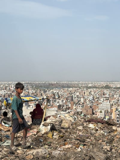 This image depicts a young boy standing in a landfill, with a densely populated cityscape in the background. The landfill is characterized by piles of garbage and debris, with a colorful tarp and makeshift shelter visible. The boy is wearing a green t-shirt and dark shorts, and appears to be barefoot or wearing sandals. In the middle ground, a person in a maroon garment is seated. The background reveals a vast urban landscape with numerous buildings of varying heights and colors, suggesting a densely populated area. This scene is characteristic of areas surrounding the Ghazipur landfill in Delhi, India, known for its large size and the significant population living and working in its vicinity.