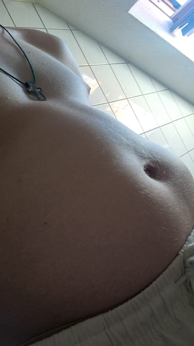 The image shows a close-up of a person's abdomen, with a focus on the navel. The person is wearing light-colored, loose-fitting pants on the left side of the frame. A necklace with a cross pendant is visible on the upper right side of the frame, resting on the person's shoulder or clavicle area. The background appears to be a tiled surface, likely a wall or floor, with light grout lines. The lighting suggests it is daytime, possibly indoors with natural light from a window or bright artificial lighting. The image is angled and possibly taken from a low perspective, looking up towards the person's torso. There are no visible signs of text in the image. The overall impression is a personal, perhaps candid, shot focusing on a specific part of the body and a piece of jewelry. Given the location context of Parchim, Germany, the scene could be within a residential or private space. The texture of the skin and the subtle variations in tone are prominent features.