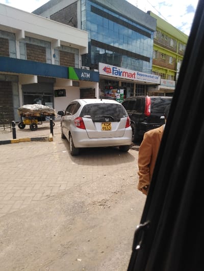 The image depicts a daytime street scene in a commercial area of Kikuyu Township ward, Kenya, viewed from inside a vehicle.

In the immediate foreground on the right, the dark frame of a vehicle's interior is visible. To the left, a tan or light brown jacket of a person is partially visible. The ground transitions from a dirt surface in the foreground to a paved area of interlocking concrete blocks towards the center-left.

A silver Honda Fit hatchback (rear view, Kenya license plate KCM 983J) is parked on the paved surface, facing away from the viewer. Behind it, a dark-colored SUV (possibly a Nissan X-Trail, identifiable by its taillight design) is partially visible, also parked. To the far left, near a yellow and black painted curb, a yellow two-wheeled cart laden with goods, covered by a light-colored tarp, is parked.

The background features several multi-story buildings. On the left, a building has a white upper facade with what appear to be bricked-up or decoratively paneled windows. Below this, a blue and green band displays "ATM" signage. The ground floor entrance features a dark roller shutter. Adjacent to this, a modern multi-story building with a dark grey upper facade and large, reflective blue-tinted glass windows dominates the center. Its ground floor prominently features a "Fairmart Supermarket Ltd" sign with an "FS" logo. To the right of the Fairmart building, a portion of a yellowish-green multi-story building with multiple windows is visible. The sky above is partially cloudy.