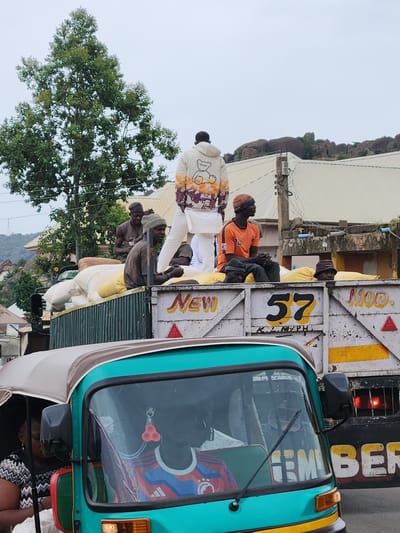 In Rukuba Road, Nigeria, a flatbed truck is loaded with numerous large, pale yellow sacks, likely agricultural produce. Several individuals are positioned atop these sacks and on the truck's railings. One man in the foreground wears an orange t-shirt and a knitted cap, while another to his right is dressed in a cream-colored hoodie adorned with a cartoon bear and orange and purple graphics, paired with white trousers. A third person, wearing a dark beanie, sits on the truck's edge to the left of the man in the orange shirt. The truck's side displays faded white paint with painted text including "NEW," "57," and "MOD." Two other men are visible behind the front figures. In the foreground, a teal-colored auto-rickshaw is partially visible, with a person in the driver's seat wearing a blue and red striped shirt. The background reveals a tree with dense green foliage and a rocky, elevated terrain with some buildings. The sky is overcast.
