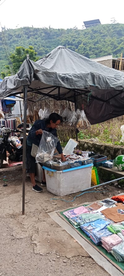 The scene shows a street vendor selling goods from a stall in Kutawaringin, Indonesia. The stall is located at the coordinates -6.974 latitude and 107.519 longitude. The vendor is a man standing in front of a large white plastic bin. He is wearing a black long-sleeved shirt and dark pants. He is holding a plastic bag in his right hand. In the bin are a variety of goods. On the left side of the image, a motorcycle is parked by the stall. There is a stack of clothing on a tarp in front of the bin. Behind the vendor, a large, gray tarp is stretched over a frame, creating a shaded space. There are some hanging bags filled with dried goods, likely fish, under the tarp. There are also several other items on a wooden table behind the vendor. There are small white plastic bags, a gray tarp, and a brown paper bag visible. The scene is located on a street with an unpaved, cracked, dirt surface. The background consists of a lush green hillside. There is a building to the right of the stall. There is a bamboo structure behind the building. The sky above is gray and overcast. The scene suggests a typical outdoor marketplace in Indonesia.