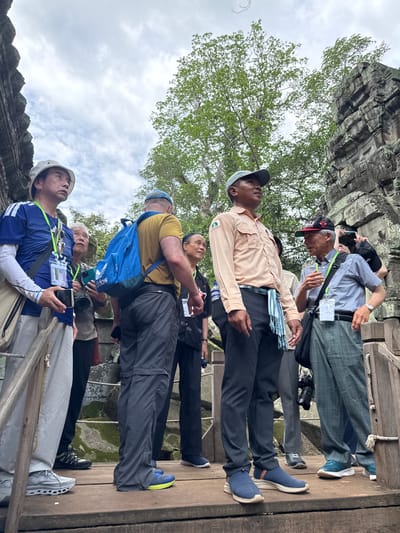 A group of individuals, likely tourists, are gathered on a wooden walkway at an ancient temple in Siem Reap, Cambodia. The temple structure, made of weathered stone, is visible in the background, partially obscured by lush green trees. The sky is overcast with scattered clouds.
In the foreground, several men are the main subjects. One man, wearing a light brown shirt and a light blue and white striped sash around his waist, stands facing slightly left, looking upwards. To his left, a man in a blue and white adidas shirt and a white hat is also looking upwards. Behind him, another man, wearing a yellow t-shirt and a blue backpack, is visible. Further to the right, a man in a blue and white striped shirt and a red-and-black baseball cap is talking, gesturing with his hand. Another man in a black t-shirt stands to the far right, partially obscured.
Several people are wearing lanyards with identification badges. Some are holding cameras and phones. The wooden walkway appears to be part of a path through the temple complex. The overall atmosphere suggests a guided tour or exploration of the historical site.