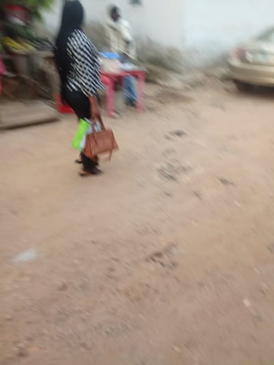 A motion-blurred image captured at ground level shows a person walking on a dirt path in Kagini, Nigeria. The individual, positioned in the left-center of the frame, is dressed in black pants and a horizontally striped black and white long-sleeved shirt. They are carrying a brown handbag in their right hand and a bright green shopping bag in their left. Behind the person, a blurred red table and another indistinct figure wearing a light-colored shirt are visible. To the far right, the rear portion of a light-colored vehicle is partially visible. The ground is composed of reddish-brown dirt and small stones. The background features a blurred white wall.