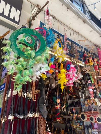 This image shows a street stall in Kathmandu, Nepal, selling a variety of handicrafts. The stall is crowded with colorful items, including dream catchers, puppets, necklaces, and bells. In the foreground, there are several dream catchers made of green yarn and feathers. Behind them, there are rows of necklaces with various pendants, some made of wood and others with religious symbols. Further back, there are several puppets dressed in traditional clothing, suspended by strings. Large drums and bells are also visible, suggesting a cultural or musical element to the stall's offerings. The stall appears to be located outdoors, possibly under an awning or overhang, with a building facade and a street visible in the background. The overall impression is one of a vibrant and bustling marketplace.
