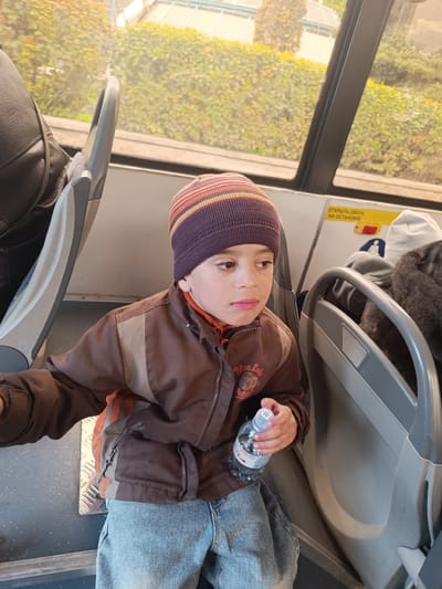 A young boy is seated inside what appears to be a public transport vehicle, possibly a bus or tram, in Simferopol, Ukraine. He is positioned on the left side of the frame, looking towards the right with a somewhat pensive or thoughtful expression.
The boy, likely between 5-8 years old, wears a striped beanie (purple, orange, and yellow/light brown stripes), a brown and beige jacket with an orange lining visible, and light blue jeans. On his jacket, a patch with the text "Blue Lake" is visible. He is holding a clear plastic water bottle in his right hand. The bottle has some Cyrillic text, including "НАТУК", and a QR code.
He is seated on a grey seat, with a part of a black backpack visible behind him on another seat to his left. To his right, another grey seat with a handle is visible, and behind it, a person wearing a grey hoodie or hat and a dark, possibly furry, coat is visible. Above these seats, a white sign with Cyrillic text reads "ОТКРЫТЬ ДВЕРЬ НА ОСТАНОВКЕ" (meaning "OPEN DOOR AT THE STOP").
Through the vehicle's windows, green foliage, likely bushes or trees, is visible, suggesting an outdoor, possibly urban or suburban, environment. The light outside appears bright but somewhat diffused, indicating a daytime scene, possibly overcast. The interior of the vehicle has a functional, utilitarian design with grey plastic seating and interior panels.