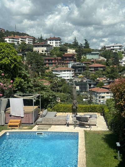 This is a high-angle, full shot of a residential area and a swimming pool. The scene is oriented vertically, with the sky at the left and the ground at the right. Several multi-story buildings with red-tiled roofs and white facades are nestled among lush green trees on a hillside. A body of water, likely a swimming pool, occupies the lower right portion of the frame. The pool is bright blue and appears to be clean and well-maintained, with a tiled edge. Adjacent to the pool is a paved area with lounge chairs and a table. The sky is partially cloudy, suggesting it is daytime, possibly late morning or early afternoon. The overall impression is one of a serene and luxurious residential setting, likely in a hilly coastal area given the view of buildings cascading down a slope. There are no people visible in the frame. The image appears to be taken from a balcony or a higher vantage point, looking down and across the landscape. The location context suggests Beşiktaş, Turkey, which is known for its hilly terrain and Bosphorus views, aligning with the visual elements in the image.