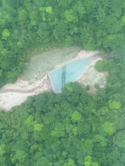 An aerial view shows a narrow river flowing through a dense forest, likely located near the city of Zhangjiajie, China. The river is mostly light blue in color and is surrounded by lush green trees on both sides. A possible dam-like structure is located towards the upper third of the river's flow, causing a pool of water to form just before the structure. There is also a bridge reflecting on the water right before the dam. The riverbed is partially visible and filled with light-colored rocks and pebbles. The photograph exhibits a slightly hazy or foggy atmosphere.