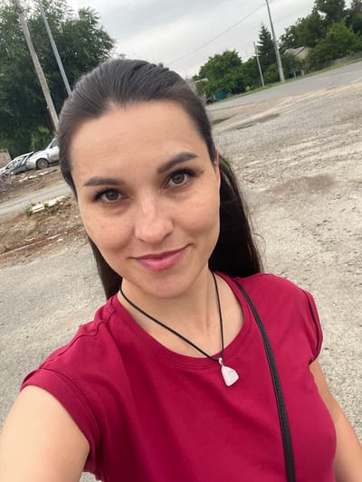 In the city of Donskoye, Russia, a close-up selfie captures a woman with dark hair, brown eyes, and light skin with freckles, smiling slightly. She is wearing a red t-shirt and a black necklace with a light-colored pendant. The background shows a gravelly area with some greenery and trees, along with a street and a few abandoned cars. The sky is overcast.