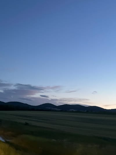The image captures a landscape scene likely near Gorna Kremena, Bulgaria, given the presence of mountainous terrain and field-like areas. The sky occupies a significant portion of the left side of the image, displaying a gradient of blue that suggests twilight or early evening. Some clouds are visible near the horizon. A mountain range is seen towards the middle of the image, its darker silhouette indicating that it is further in the distance. The right side of the image shows a field or grassland that appears to be relatively flat, and green. The photo seems to be taken at dusk or dawn. The image is slightly out of focus.