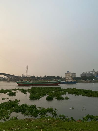 The image depicts a wide river with several boats and industrial structures along the bank. In the foreground, thick patches of green aquatic plants float on the water's surface. Two cargo ships are visible; one, larger and red and green, is moving from left to right in the middle ground. A smaller, blue and white vessel is positioned further to the right, behind the first ship.
In the background, a large bridge spans the river on the left side of the frame. Behind the bridge and river, a cityscape emerges, characterized by tall buildings and industrial facilities, including silos. A prominent lattice tower, likely a power transmission pylon, stands out against the hazy sky. The sky is overcast and appears to be either early morning or late afternoon, with a soft, diffused light. The overall impression is of an active industrial waterway in Tarabo, Bangladesh.