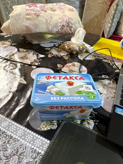 This is a close-up, eye-level shot of a table covered with a black and white floral-patterned cloth. A blue plastic container of feta cheese, labeled "FETAXA" in red Cyrillic lettering, is the prominent subject in the foreground. The container is partially open, revealing white cheese cubes and a small red tomato on top. A green leaf, likely basil, is also visible. Text on the container indicates "Без рассола" (without brine) and "ДЕРЖИТ ФОРМУ" (holds its shape). To the right, a portion of a laptop with an "Intel Core i3" sticker is visible, along with a black cable. In the background, stacked fabrics with floral patterns and lace trim are piled up, as well as a plastic bag containing small, yellow, spherical objects. The overall scene is indoors, and no specific location in Bukhara, Uzbekistan, can be determined from the visual information provided. There are no people or actions depicted.