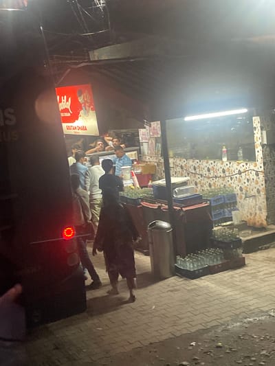 The image captures an outdoor scene at night in Katauhar Kalan, India, likely near a food stall or dhaba. In the foreground, a dark vehicle with a lit red tail light is visible on the left. A person's hand is partially visible in the bottom left corner, obscuring part of the vehicle. The main focus of the image is a bustling stall with several individuals. Above the stall, a bright red advertisement for "LAL ICE CREAM" is displayed, with the text "GOUTAM DHABA" below it.

Inside and around the stall, numerous people are present. Some individuals appear to be staff, possibly serving customers or preparing food, while others seem to be patrons. They are dressed in casual clothing. Behind the counter, a selection of beverages and food items are visible, including crates of bottled drinks, particularly on the right side of the stall, stacked on the ground. A trash bin is positioned to the left of the stall. The ground is paved with interlocking bricks, and there is some wetness or debris on the ground, suggesting it might be an area with foot traffic and potential spills. The overall ambiance suggests a lively, late-night establishment.
