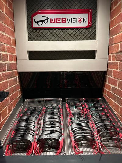 The image depicts a glasses dispensing or storage unit located in Chessy, France. A red-brown brick wall flanks both sides of a built-in structure. The upper section features a light-colored frame surrounding a dark metal grid, on which a red and white sign displays "WEBVISION" alongside a black stylized glasses icon and a camera shutter icon. Below this, a dark, reflective surface occupies the mid-section. The lower half of the unit contains multiple rows of specialized glasses, each with a black frame and prominent red earpieces, meticulously arranged in silver-colored mesh trays. Approximately four such trays are visible, uniformly populated with the glasses. No individuals are present in the scene.