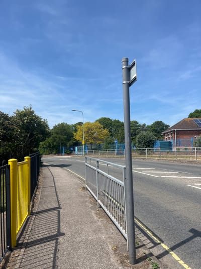 A photograph shows an outdoor scene, possibly a street in East Suffolk, United Kingdom, on a sunny day with a clear blue sky. The camera is positioned relatively low to the ground, and the image is rotated to the left. A gray metal pole with a rectangular object on top is in the foreground, extending horizontally across the upper part of the frame. Attached to the pole is a gray metal railing. A paved road with white and yellow lines runs parallel to the railing. Beyond the road and railing are buildings, trees, and a blue fence or structure. A yellow structure runs on the bottom of the photo with its shadow covering the sidewalk.