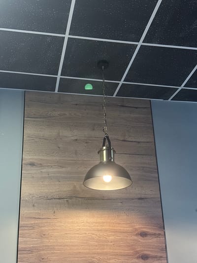 The image shows a modern interior scene featuring a hanging lamp with a metal chain and a brushed metal shade. The lamp is illuminated by a warm light, casting a soft glow on the wooden paneling behind it. The wall to the left is decorated with a grid pattern of dark tiles interspersed with white lines, creating a textured and contemporary look. A faint green circular light is visible on this tiled wall, possibly a safety marker or indicator. The overall setting appears to be a well-lit indoor space, possibly a restaurant, cafe, or modern living area, in Murino, Russia. There is no visible text in the image. The lighting suggests it could be evening or indoors with artificial light. The scene conveys a sense of modern design and ambiance.