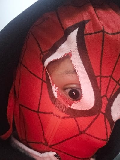 A close-up shows a person wearing a red Spider-Man mask with black web designs. Only one eye is visible through a hole in the mask, peering out. The mask stretches over the face, and the person is wearing a black hooded jacket. The hood is up, framing the mask. Part of a white garment peeks out near the zipper of the jacket. The lighting is soft, highlighting the texture of the mask.