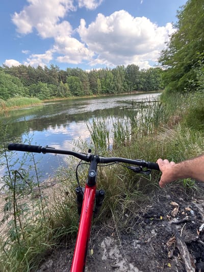The media file shows a first-person perspective of someone holding the handlebars of a red bicycle. The bicycle is positioned on a path next to a body of water, likely a lake or river, with lush green trees and vegetation surrounding it. The water reflects the blue sky and white, fluffy clouds. The ground next to the path appears to be dark soil with some debris, possibly from a fire pit, and dry grass.
The scene suggests an outdoor activity, likely cycling, in a natural setting. The sunlight indicates it is daytime, and the presence of clouds suggests potentially variable weather. The dense greenery and trees point to a location with abundant plant life. The visible text on the bicycle frame reads "XTC". The overall impression is one of a scenic outdoor excursion.