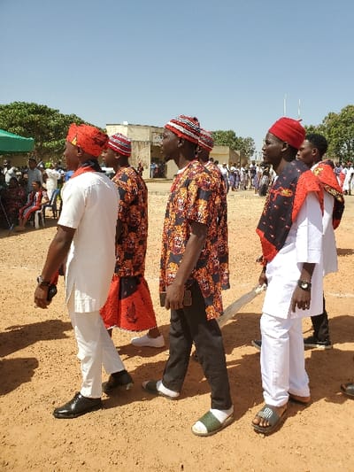 The image displays an outdoor scene in Bar Gada, Nigeria, under a clear blue sky and bright daylight. The foreground features five men walking from left to right across a dry, light brown dirt ground. The leftmost man, partially visible, wears a white tunic, white trousers, black dress shoes, and a red patterned cap, holding a dark object in his left hand. The second man wears a black short-sleeved shirt with orange and red patterns and a red and white striped cap. The third man, centrally located, wears a similar black and orange patterned short-sleeved shirt, dark trousers, a red and white striped cap, white socks, and olive green sandals, holding a dark object in his right hand. The fourth man wears a white tunic and trousers, a red cap, dark sandals, and has a black and red patterned fabric draped over his left shoulder, carrying a light-colored staff in his right hand. The fifth man, partially visible, wears a white tunic. In the left mid-ground, multiple individuals are seated on white plastic chairs under a green canopy. Behind them, a light-colored, multi-story building with windows is visible. Trees and additional individuals are present in the background, to the left and right of the building. Two thin poles are observable in the far right background.