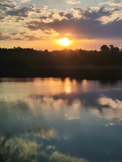 The image depicts a sunset scene over a body of water. The sun is partially obscured by clouds, with visible sun rays emanating from behind them. The water reflects the colors of the sky and clouds, showing a mix of orange, yellow, and grey hues. A dark silhouette of trees and vegetation is visible on the horizon, separating the sky and the water. The location is Ermolino, Russia.