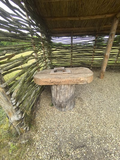 The image depicts an outdoor scene within a rustic structure that appears to be made from natural materials. The structure has a thatched roof supported by wooden beams, and its walls are constructed from horizontally woven branches. In the center of the structure, a rough-hewn wooden table stands on a thick, log-like base. The table top is wide and flat, with a visible seam or crack. On the table's surface, there are two small, rectangular wooden objects. The ground within the structure is covered with small, light-colored gravel. To the left of the structure, a grassy area is visible, with some dry grass and foliage around the base of the structure. The background shows a green, grassy field with a line of trees or vegetation in the distance, and the sky is overcast. The scene suggests an outdoor seating area or a primitive workstation, possibly in a rural or historical setting. The location is specified as Камянюцкі сельскі Савет, Belarus.