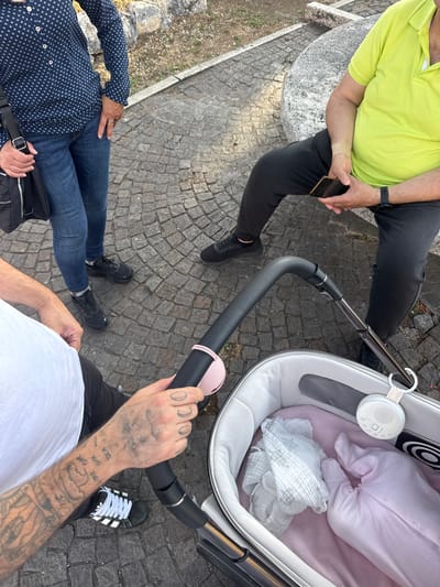 The image shows an overhead shot of three people and a baby stroller.
A person wearing a green polo shirt and black pants sits on a stone structure, holding a phone. Another person, dressed in a blue patterned shirt and jeans, sits nearby. A third person, wearing a white t-shirt and black pants, stands holding the handle of a baby stroller. The stroller contains a baby wrapped in pink clothing and a white blanket. A white noise machine hangs from the stroller.
The setting appears to be an outdoor public area, with cobblestone ground. The location is Aprilia, Italy.
