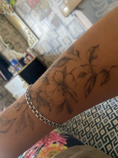 The image provides a close-up view of a person's light brown arm, adorned with a floral tattoo and a silver chain bracelet. The arm has fine hair and healthy skin. The tattoo is rendered in black and grey, depicting a winding branch or vine with multiple blooming flowers and an array of leaves, covering a significant portion of the forearm. The design features both distinct linework and subtle shading. A chunky silver bracelet with interlocking oval links is worn on the wrist, partially covering the lower section of the tattoo.

The background suggests an indoor setting, likely a domestic interior in Topoli, Bulgaria. In the upper right, there is a patterned fabric, possibly a cushion or upholstery, with a repeating blue and white geometric design. Adjacent to it, on the right side of the frame, is a brightly colored fabric featuring large, abstract floral patterns in shades of pink, yellow, and green. The lower left and bottom areas of the image show a reflective surface, possibly a polished floor or a large mirror, which reflects parts of the room. This reflection reveals what appears to be a patterned floor or rug, and ornate furniture with gold accents. Blurred figures and objects, including a blue item, are faintly visible in the deeper background reflections.

No specific activity is taking place; the arm appears stationary. The lighting indicates an indoor environment, likely illuminated by ambient light. There is no visible text in the image.