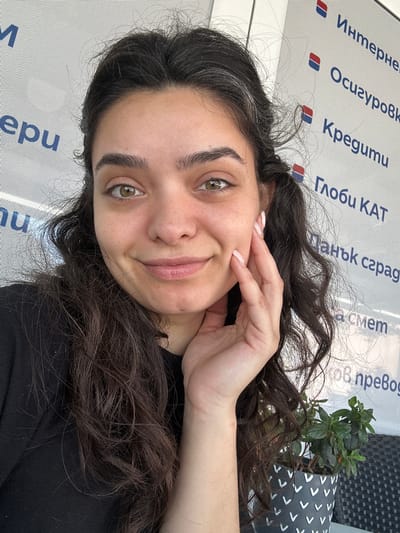 The image is a medium shot selfie of a young woman with dark, curly hair and light-colored eyes. She is looking at the camera and smiling slightly, with her right hand resting on her cheek. She is wearing a black top. The background appears to be an outdoor setting with a white textured wall that has blue lettering on it, possibly indicating a business or establishment. A small potted plant with green leaves is visible to her right. The lettering on the wall includes what appears to be Bulgarian text, with phrases like "Интерне", "Осигуровк", "Кредити", and "Папк сград". This suggests the location is likely in Bulgaria, and based on the provided context, specifically Malevo.