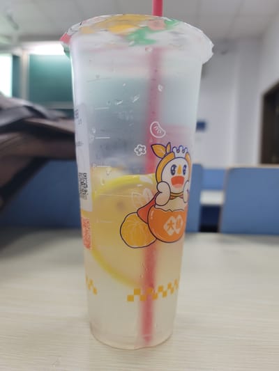 A clear plastic cup with a lid sits horizontally on a light-colored, possibly wooden surface. The cup is filled with a pale yellow liquid and has a red straw. Cartoon graphics decorate the cup's exterior. Condensation droplets cling to the cup. A blurred background suggests an indoor setting, possibly a room within a building. Elements visible in the background include a chair and part of a window. The photo was most likely taken in Jiangning District, China.