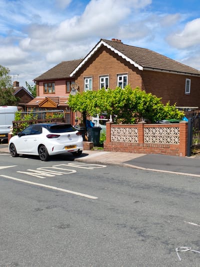 In Sandwell, United Kingdom, on a sunny day with scattered clouds, a white Vauxhall Corsa is parked on the left side of a residential street. The car, featuring a black roof and tinted windows, has a yellow license plate with the registration "SXIIMAH". A speed bump is visible on the asphalt road in the foreground.
Behind the car, a brick house with a brown tiled roof and white decorative trim stands. The house has multiple windows, some with white frames and others with brown frames. A porch area with wooden supports is attached to the front. Adjacent to the house, a low brick wall with decorative concrete panels separates the property from the street. Green trees and foliage grow behind the wall and around the house. Two dark green wheelie bins are positioned near the property's entrance.
To the left of the house, part of a white van is visible, with a license plate that reads "ZI XYP". The environment appears to be a suburban residential area.
