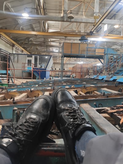 The image depicts a first-person perspective of a person's legs and feet, clad in black work boots and grey socks, resting on machinery within an industrial facility. The background reveals a large, open-plan workshop with a high ceiling supported by metal beams and industrial lighting fixtures. Multiple conveyor belts and metal structures are visible, suggesting a manufacturing or processing environment, possibly related to woodworking or material handling given the presence of wood scraps on the belts. In the middle ground, a control booth with windows is situated above the machinery. The overall setting is an indoor industrial space, with a focus on the equipment and the immediate foreground occupied by the person's footwear.
