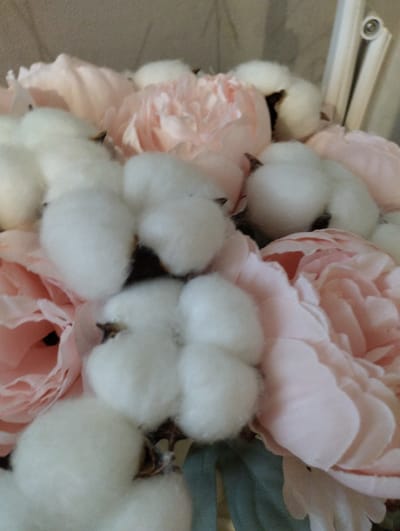 A close-up, angled shot showcases a bouquet of light pink peonies and cotton bolls. The peonies have soft, layered petals that range from pale to slightly deeper pink. Nestled among the flowers are several cotton bolls, their fluffy white fibers contrasting with the delicate petals. Some bolls reveal dark brown centers where they were attached. The background is a textured, light beige surface, possibly a wall or fabric. A white, cylindrical object with a small circular detail is partially visible in the upper right corner, suggesting the bouquet might be placed near a window or on a windowsill. The lighting is soft, casting gentle shadows that highlight the textures of the flowers and cotton. The scene appears to be an indoor arrangement, with no discernible details about its specific location within Щучье, Russia.