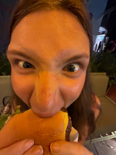 The image shows a close-up, sideways view of a person's face as they are about to take a bite out of a cheeseburger. The person's eyes are wide open, and their mouth is open, with the burger positioned near their lips. The burger appears to have a beef patty and melted cheese. The background is out of focus but suggests an outdoor setting at night, possibly a street or dining area, with some greenery and lights visible. The person has brown hair. The overall impression is a spontaneous moment captured while eating. The location context is Shanghai, China. No visible text is present in the image.