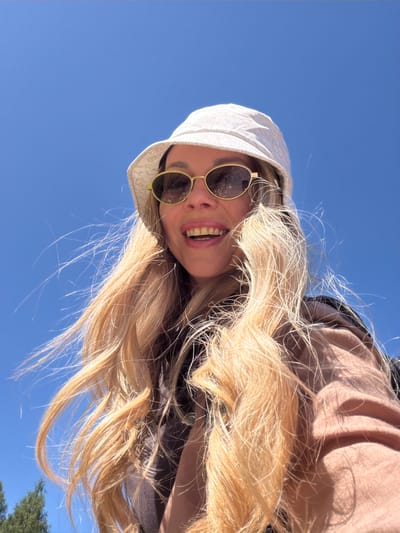 A low-angle, close-up shot shows a woman in a white bucket hat and gold-rimmed oval sunglasses against a bright blue sky. Her long blonde hair is windblown. She is smiling, revealing her teeth. She is wearing a brown jacket over a tan top. The image does not provide specific details to determine the location within Sauris di Sopra / Plozn / Sauris Disore, Italy.